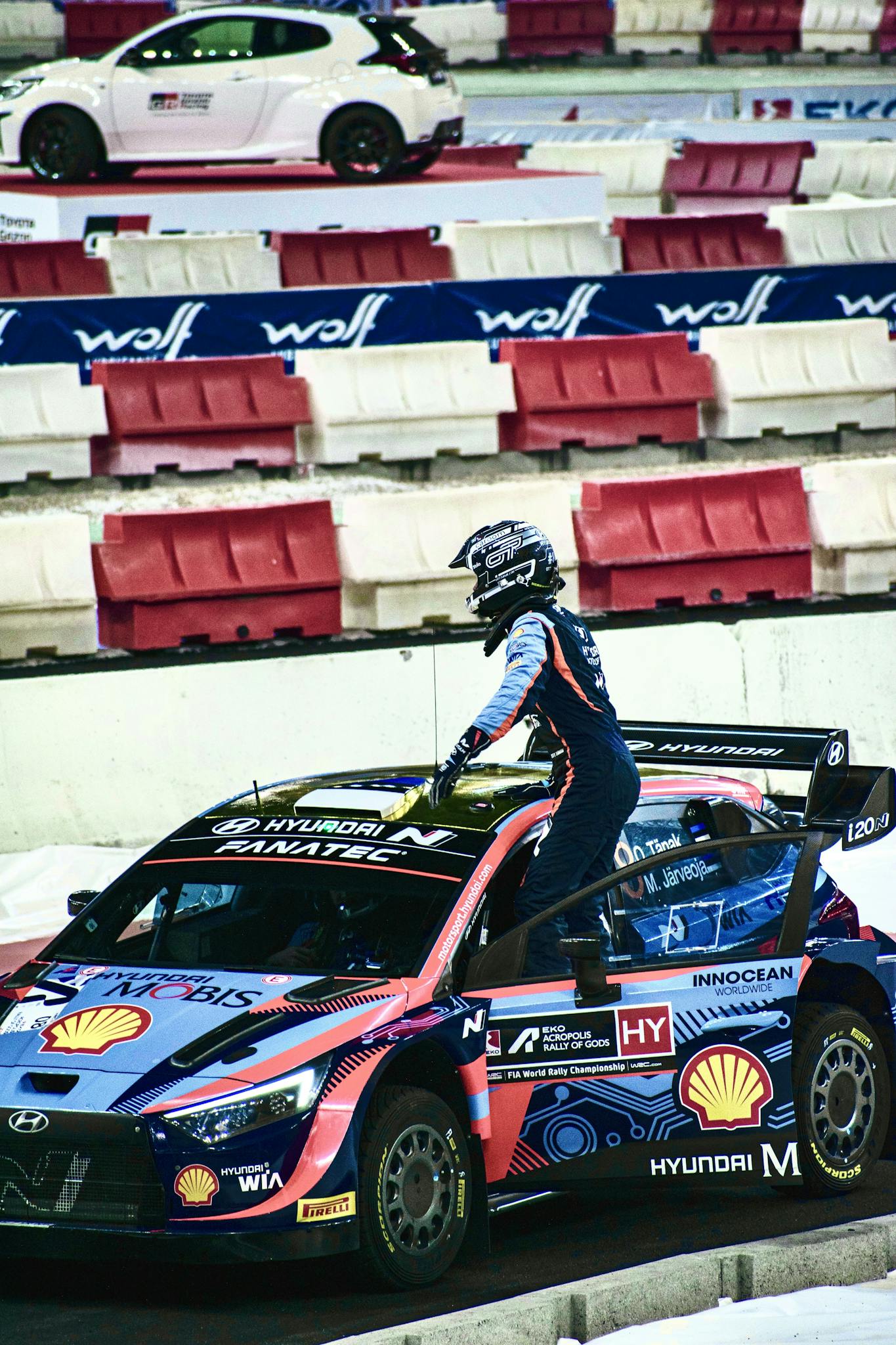 A rally driver in sports gear exits a Hyundai race car on a racetrack, showcasing motorsport action.